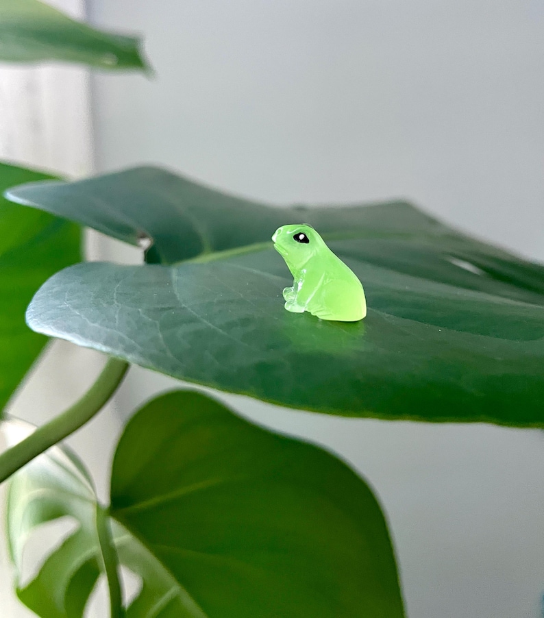 May include: A small green glass frog figurine sitting on a large green leaf.
