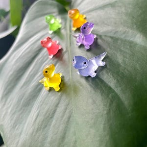 May include: A set of six small, colorful, translucent figurines shaped like dinosaurs. The figurines are arranged in a circle on a green leaf. The colors of the figurines are green, yellow, red, purple, blue, and light blue.