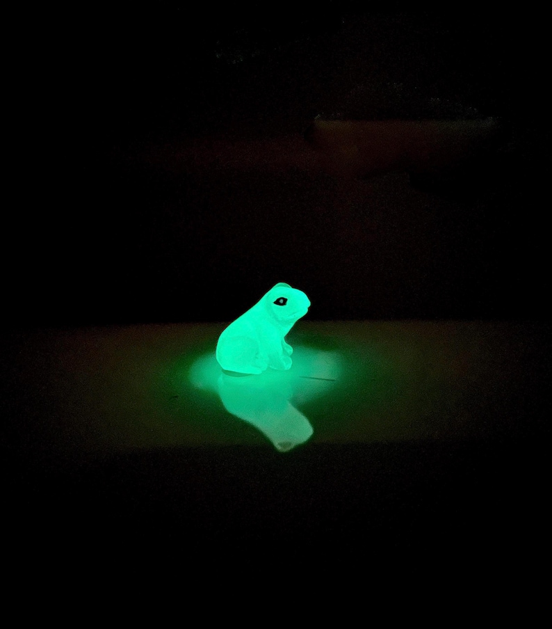 May include: A small, green, glow-in-the-dark frog figurine. The frog is sitting on a white surface and is glowing brightly.