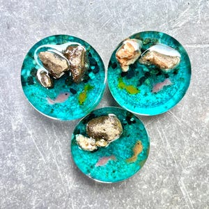 May include: Three round resin art pieces with a teal blue background, featuring small rocks, pink and yellow fish, and a white accent. The pieces are arranged on a gray surface.