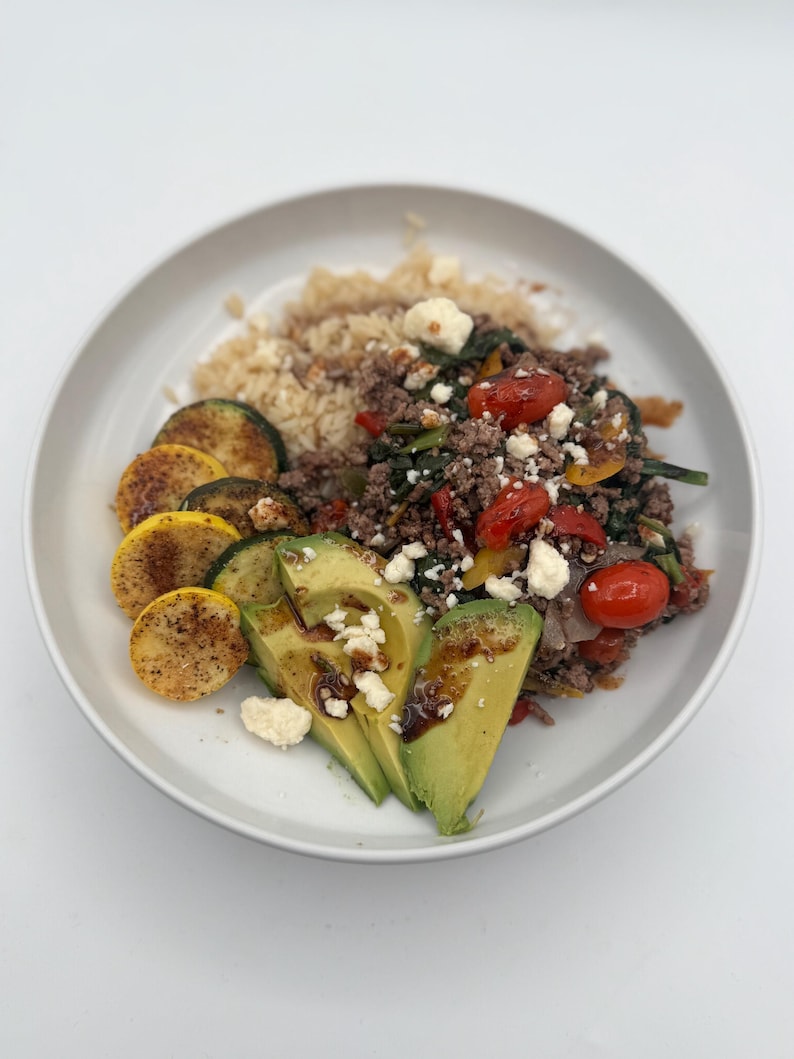 May include: A white bowl filled with a meal of brown rice, ground beef, spinach, tomatoes, zucchini, avocado, and feta cheese.