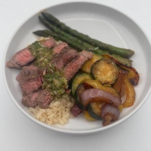 May include: A white bowl with a serving of brown rice, grilled steak with a green sauce, asparagus, and sauteed vegetables including zucchini, red onion, and yellow bell peppers.