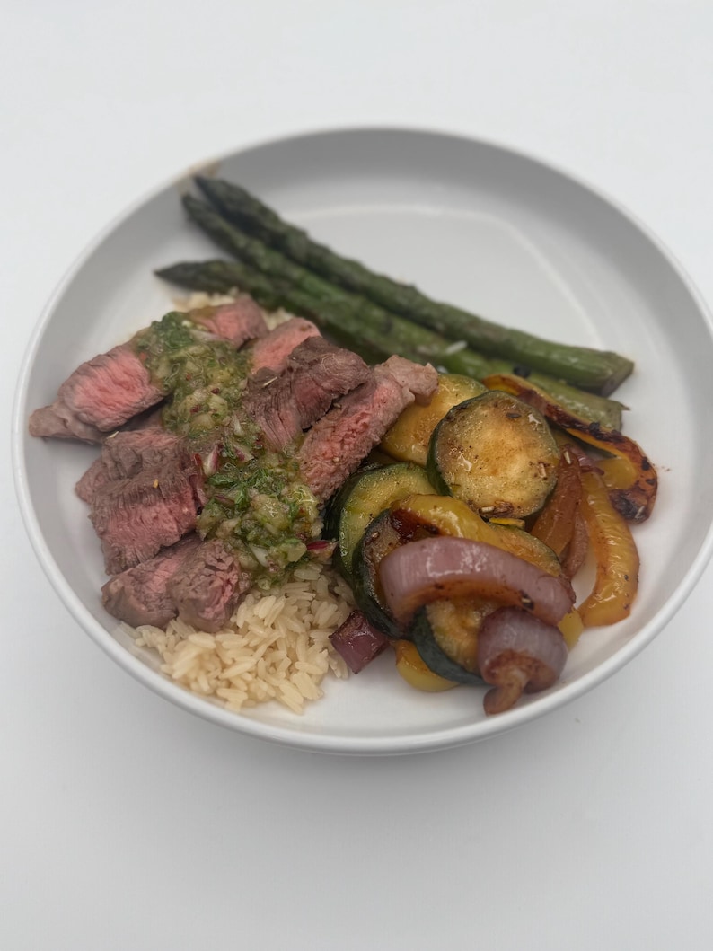 May include: A white bowl with a serving of brown rice, grilled steak with a green sauce, asparagus, and sauteed vegetables including zucchini, red onion, and yellow bell peppers.