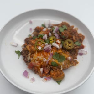 May include: A white plate with a serving of ground beef casserole topped with shredded cheese, diced red onion, sliced jalapeno peppers, and chopped cilantro.