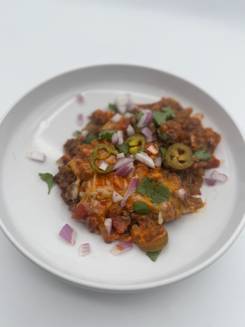 May include: A white plate with a serving of ground beef casserole topped with shredded cheese, diced red onion, sliced jalapeno peppers, and chopped cilantro.