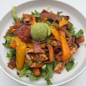 May include: A white bowl filled with a colorful salad featuring chicken, peppers, onions, sweet potatoes, and a dollop of guacamole.