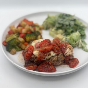May include: A white plate with a chicken breast topped with mozzarella cheese and roasted cherry tomatoes. The chicken breast is surrounded by roasted vegetables and a side salad.