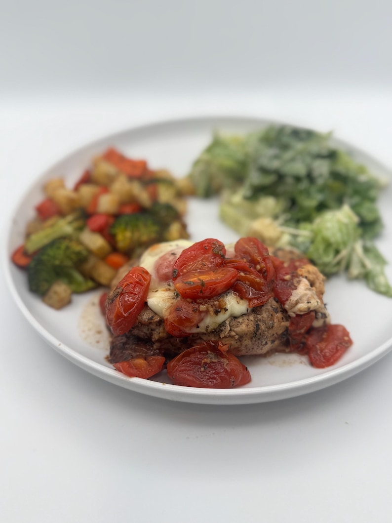May include: A white plate with a chicken breast topped with mozzarella cheese and roasted cherry tomatoes. The chicken breast is surrounded by roasted vegetables and a side salad.