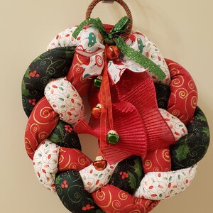 May include: A braided Christmas wreath with red, green, and white fabric. The wreath is decorated with a red and green bow, a red and white ribbon, and jingle bells.