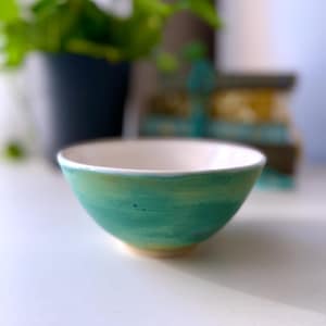 May include: A small, white ceramic bowl with a green and yellow glaze. The bowl has a rounded shape and a slightly flared rim.