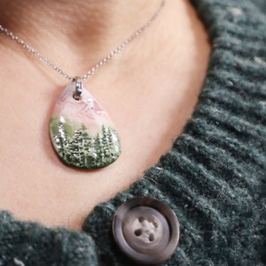 May include: A silver chain necklace with a teardrop-shaped pendant featuring a hand-painted landscape scene of a mountain range with pink and green hues.