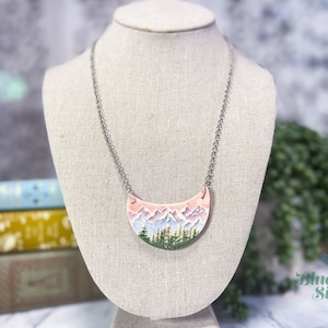 May include: A silver chain necklace with a crescent-shaped pendant featuring a painted landscape of mountains, trees, and a pink and purple sky.