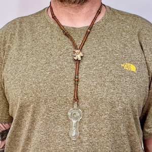 May include: A brown cord necklace with a glass pipe and a wooden bear pendant. The necklace is worn by a person wearing a green shirt with a logo that says "The North Face".