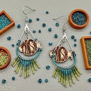 May include: A pair of dangle earrings featuring a central laser-cut wooden fish shape suspended within a silver-toned metal teardrop frame. The lower portion of each frame is adorned with a fringe of small glass seed beads in shades of teal, blue, and lime green.