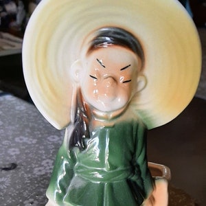 May include: A ceramic planter shaped like a person wearing a wide-brimmed hat and holding a bucket. The planter is painted in shades of green, brown, and white.