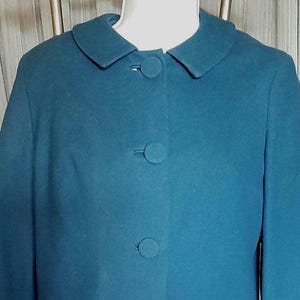 Puede incluir: Traje de dos piezas color teal. La chaqueta tiene cuello redondo, tres botones grandes y mangas tres cuartos. La falda llega a la rodilla. El traje está hecho de una tela lisa y uniforme. El traje se exhibe en un maniquí.