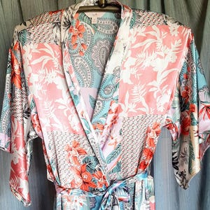 May include: A colorful floral print robe with a tie waist. The robe features a patchwork design with pink, blue, and white floral and paisley patterns. The sleeves are wide and the robe is hanging on a rack.