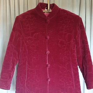Peut inclure: Veste en velours bordeaux avec col officier et manches longues. La veste présente un motif floral et se ferme par quatre boutons assortis sur le devant. La veste est suspendue à un cintre doré.
