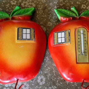 以下が含まれることがあります： 2つの赤いリンゴの形をした温度計。それぞれに黄色のミミズと窓がついています。温度計には、金色の枠で囲まれた白黒の窓と、金色の枠で囲まれた白黒の温度計があります。ミミズは黒い蝶ネクタイと黒いシルクハットをしています。