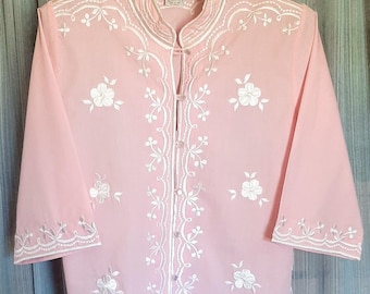 May include: A pink button-up blouse with white embroidery of flowers and vines. The blouse has a mandarin collar and three-quarter sleeves.