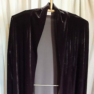 May include: A long, open-front, dark purple velvet jacket. The jacket has long sleeves and a draped collar. It is hanging on a metal clothing rack in front of a white curtain.