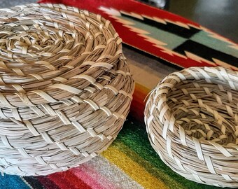 May include: Two handwoven baskets. One is a round basket with a lid, and the other is a smaller, open basket. Both are made of natural-colored woven material with white accents. The baskets are on a colorful patterned textile.