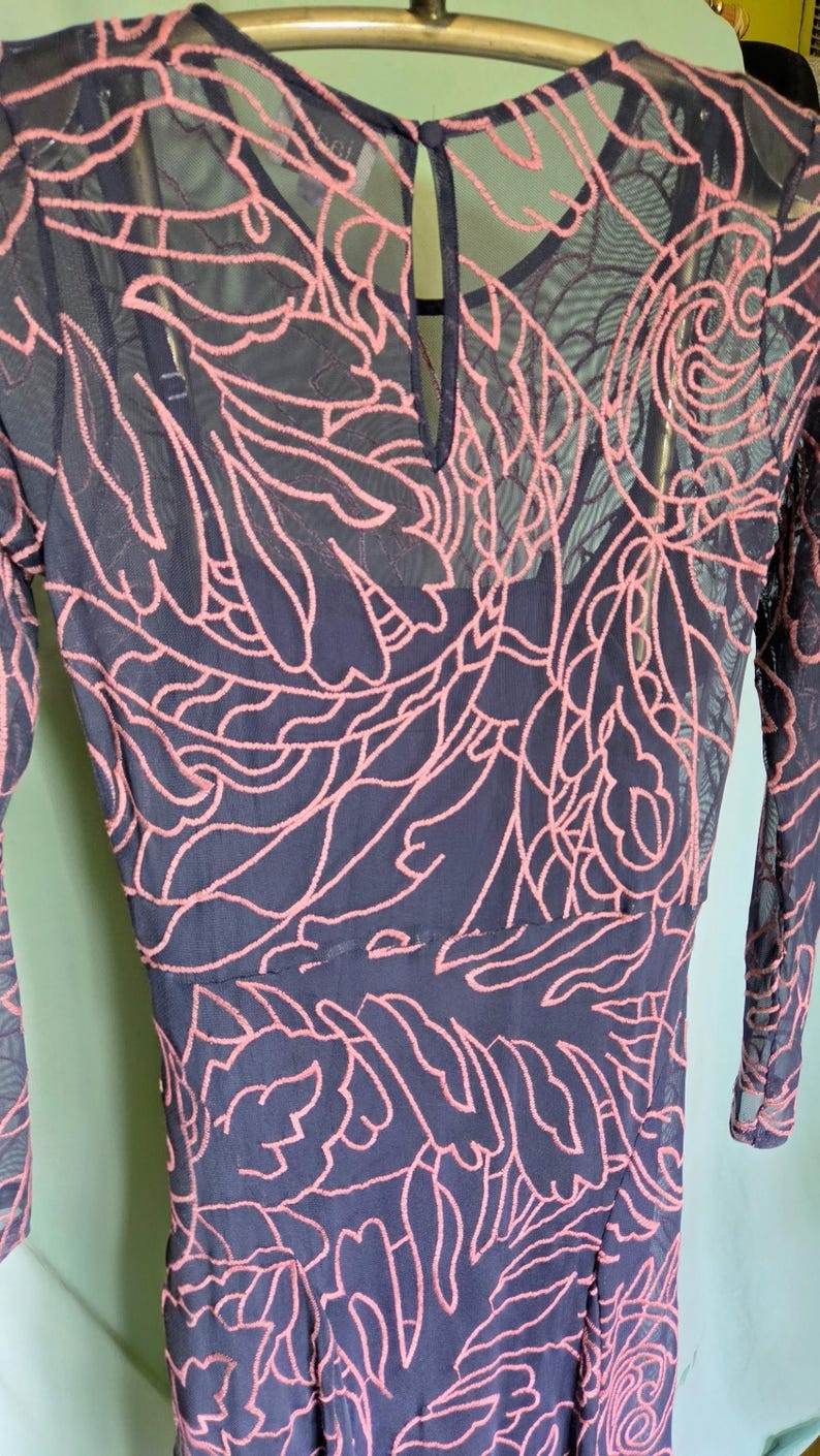 May include: A navy blue dress with long sleeves and a keyhole back. The dress features a pink floral embroidery design over a sheer mesh overlay. The embroidery creates a detailed, textured pattern across the garment.