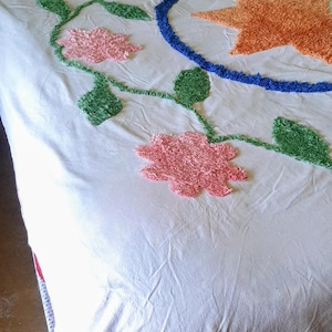 May include: White bedspread with embroidered floral and star designs. The flowers are pink, the leaves are green, and the star is orange. A blue circle surrounds the star. The headboard is made of woven bamboo.