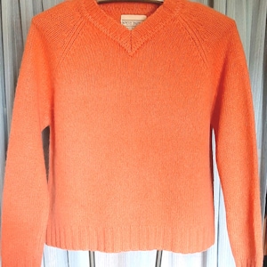 May include: A vibrant orange V-neck sweater. The sweater has long sleeves and a ribbed hem and cuffs. The neckline is a classic V-shape. The sweater is made of a soft, textured material. A small tag is visible near the neckline.