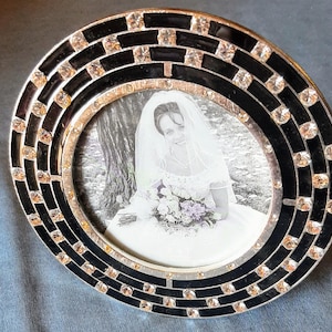 Puede incluir: Un marco de fotos negro y plateado con un borde decorativo de diamantes de imitación. El marco contiene una foto en blanco y negro de una novia con un vestido de novia sosteniendo un ramo de flores.