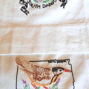 Könnte beinhalten: Bestickter weißer Stoff mit zwei Designs. Das obere Design zeigt lila Blumen und die Worte "South Dakota". Das untere Design zeigt einen Vogel innerhalb der Umrisse des Staates mit den Worten "South Dakota".