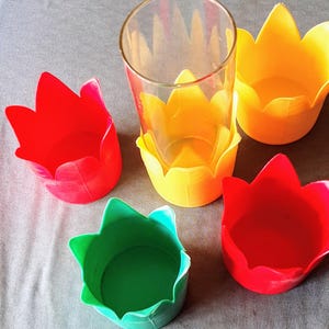 May include: A selection of brightly coloured, tulip-shaped containers in red, yellow, and green. A clear glass sits inside a yellow container. The containers are arranged on a grey surface, creating a vibrant display of colours and shapes.