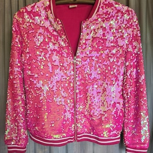 May include: A bright pink sequined bomber jacket with a full zipper closure. The jacket has a ribbed collar, cuffs, and hem with white and red stripes. The jacket is hanging on a wooden hanger.