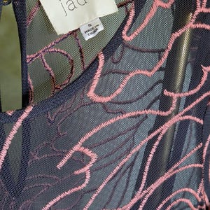 May include: Close-up of a navy blue dress with pink floral embroidery. The dress has a sheer overlay, and the tag reads "jade" with "Made in China" printed on it. The dress is size small.