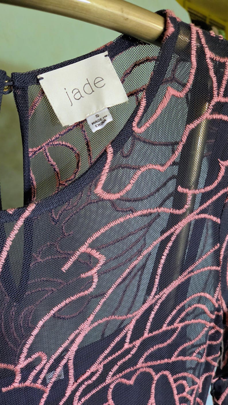 May include: Close-up of a navy blue dress with pink floral embroidery. The dress has a sheer overlay, and the tag reads "jade" with "Made in China" printed on it. The dress is size small.