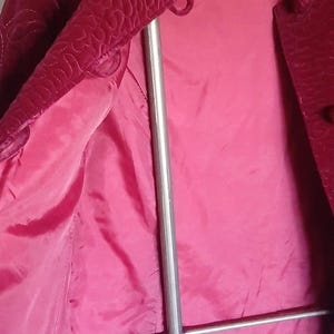 Puede incluir: Chaqueta de terciopelo burdeos con dise&ntilde;o acolchado en el cuello y mangas superiores. El forro interior de la chaqueta es de color rosa brillante. La chaqueta est&aacute; colgada en una percha de metal.
