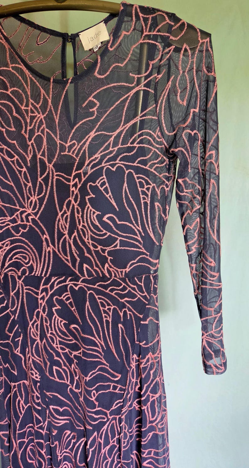May include: A navy blue dress with a sheer overlay featuring a pink floral pattern. The dress has long sleeves and a round neckline. The brand name "jade" is visible on a tag near the neckline. The dress is a fit and flare style.