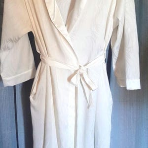 May include: A white, long-sleeved robe with a shawl collar and a matching tie belt. The robe has a subtle, textured pattern and pockets. The sleeves have a cuff detail. The robe is hanging against a blue background.