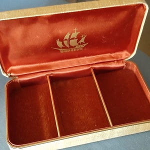 Könnte beinhalten: Geöffnete Vintage-Schmuckschatulle mit cremefarbenem Äußeren und rotem Samt-Innenraum. Das Innere hat drei Fächer und ein goldenes Schiffsemblem auf dem Deckel. Die Box ist rechteckig und hat einen Klappdeckel.