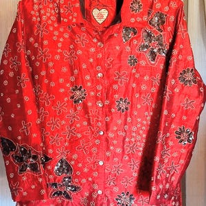 Puede incluir: Una camisa roja brillante con botones, un estampado floral y adornos de lentejuelas. La camisa tiene cuello y mangas largas. Un parche en forma de corazón está en el cuello. La camisa está colgada en una percha de madera.