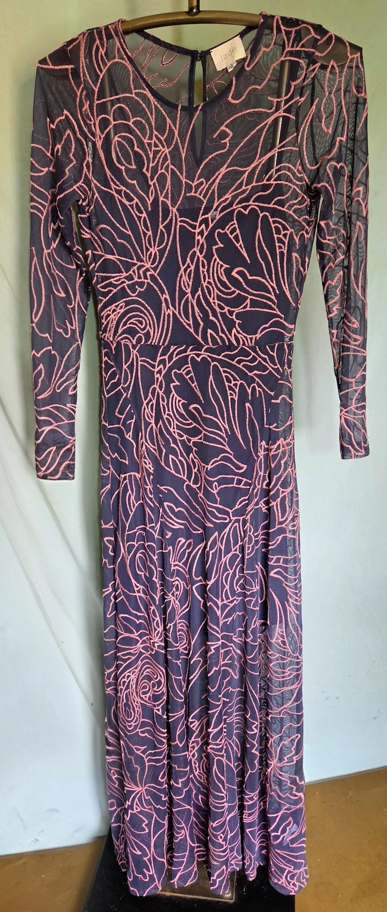 May include: A navy blue maxi dress with a sheer pink floral pattern overlay. The dress has long sleeves and a scoop neck. The label reads "jade".