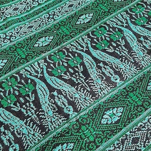 May include: Close-up of a woven textile with a repeating pattern of peacocks and floral motifs in shades of green, teal, and black. The design features diagonal stripes and intricate details.