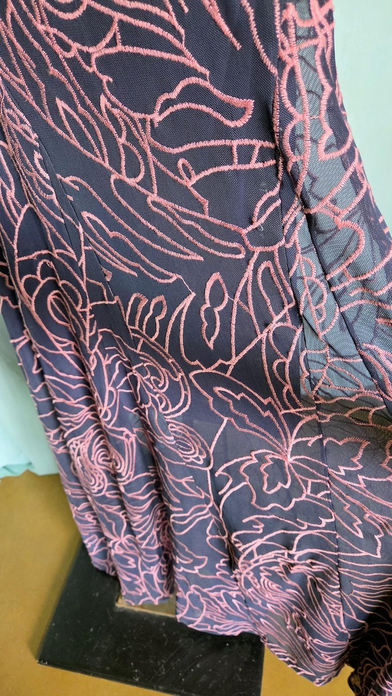 May include: Close-up of a navy blue dress with a floral pattern embroidered in a light pink colour. The dress has a sheer overlay, and the embroidery creates a textured effect.