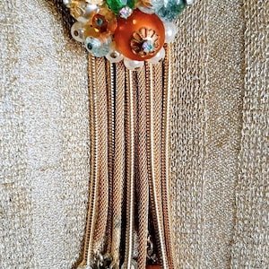 May include: Gold-toned necklace with a cluster of faux pearls, orange and green beads, and gold leaf accents. The necklace features multiple strands of gold chain that cascade down, ending with dangling beads.