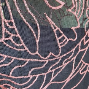May include: Close-up of a black mesh fabric with a pink embroidered floral design. The intricate pattern features flowing lines and organic shapes, creating a delicate and textured appearance. The design is visible against a dark background.
