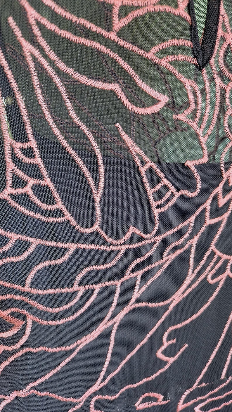 May include: Close-up of a black mesh fabric with a pink embroidered floral design. The intricate pattern features flowing lines and organic shapes, creating a delicate and textured appearance. The design is visible against a dark background.