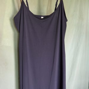 May include: A dark purple slip dress with thin spaghetti straps. The dress is hanging on a brass-coloured hanger against a light green background. The dress has a simple, flowing design.