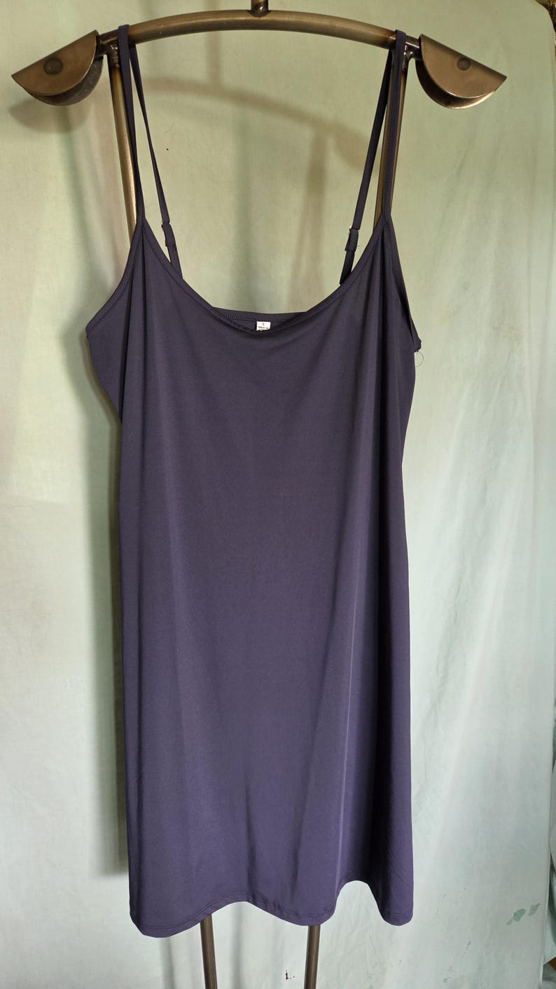 May include: A dark purple slip dress with thin spaghetti straps. The dress is hanging on a brass-coloured hanger against a light green background. The dress has a simple, flowing design.