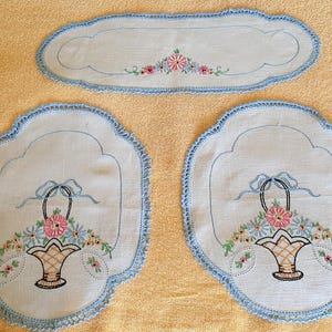 May include: A set of three embroidered linen pieces. Two are placemat-shaped with a floral basket design and scalloped blue edges. The third is a smaller, oblong piece with a floral design and blue trim. The embroidery features pink, blue, and green flowers.
