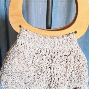 Op de afbeelding: Een beige, handgemaakte gehaakte tas met een houten handvat. De tas heeft een cirkelvormig houten handvat en een gedetailleerd, getextureerd haakpatroon. Het ontwerp van de tas is neutraal van kleur, geschikt voor diverse outfits.